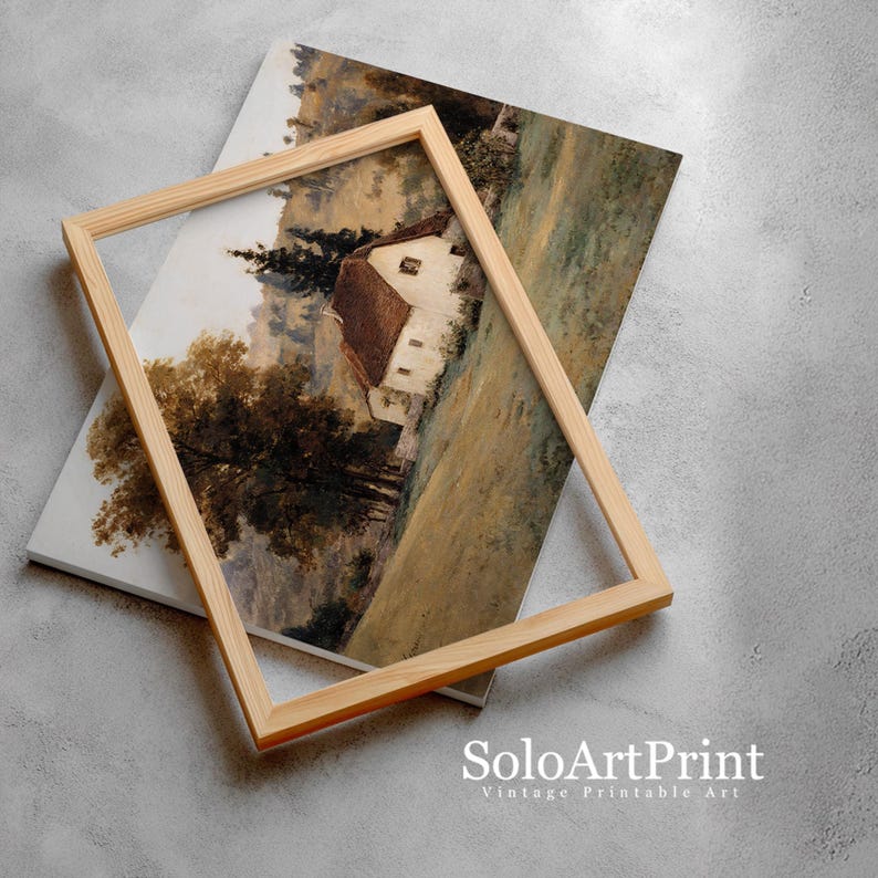 May include: A rectangular light-colored wooden frame rests on top of a landscape art print. The print depicts a white cottage with a brown roof nestled in a field with tall trees and rolling hills in muted earth tones of brown, green, and beige.