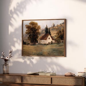 May include: A framed landscape painting depicting a white cottage with a brown roof nestled in a grassy field. A large tree with golden-brown foliage stands to the left, while rolling hills and evergreen trees appear in the background.