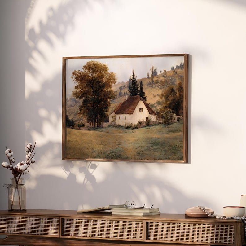 May include: A framed landscape painting depicting a white cottage with a brown roof nestled in a grassy field. A large tree with golden-brown foliage stands to the left, while rolling hills and evergreen trees appear in the background.