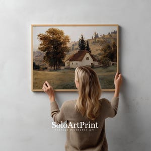 May include: A rectangular landscape art print in a light wood frame featuring a white cottage with a brown roof nestled in a grassy field with large trees and rolling hills in the background. The text SoloArtPrint is visible at the bottom.
