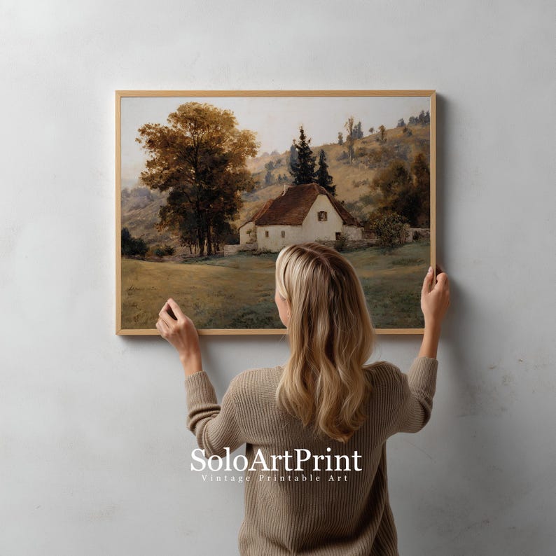 May include: A rectangular landscape art print in a light wood frame featuring a white cottage with a brown roof nestled in a grassy field with large trees and rolling hills in the background. The text SoloArtPrint is visible at the bottom.