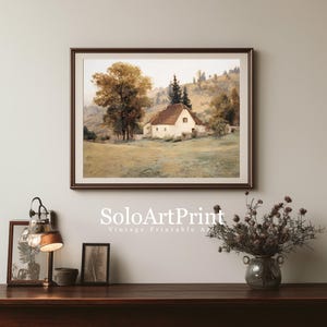 May include: A framed landscape painting featuring a white cottage with a brown roof nestled in a grassy field with large trees and rolling hills in the background. The artwork is displayed on a wall above a wooden surface with a lamp and a vase.