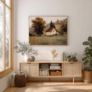 May include: A rectangular landscape painting in a thin wooden frame depicting a white cottage with a brown roof nestled in a grassy field with large trees and rolling hills in the background, rendered in muted earth tones of brown, green, and cream.