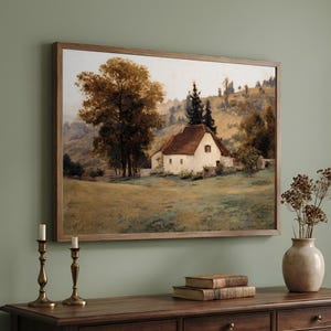May include: A framed landscape painting depicting a white cottage with a brown roof nestled in a grassy field, surrounded by large trees and rolling hills in muted earth tones of brown, green, and cream, displayed in a wooden frame.
