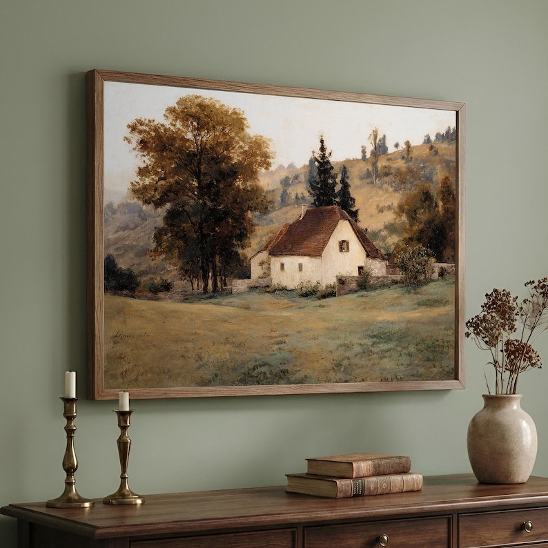 May include: A framed landscape painting depicting a white cottage with a brown roof nestled in a grassy field, surrounded by large trees and rolling hills in muted earth tones of brown, green, and cream, displayed in a wooden frame.