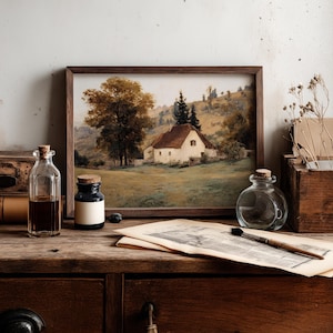 May include: A framed landscape painting depicting a white cottage with a brown roof nestled in a grassy field with large trees and rolling hills. The artwork is displayed alongside glass bottles, a paintbrush, and aged paper on a wooden surface.