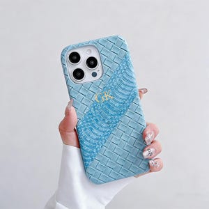 May include: A light blue phone case with a woven pattern and a textured, snake-like design. The case has a gold monogram and a camera cutout. The phone case is held in a hand.