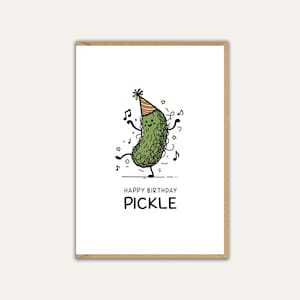May include: A white birthday card featuring a cartoon pickle in a party hat, dancing amidst musical notes and stars. The words "HAPPY BIRTHDAY PICKLE" are printed in black below the illustration. The card is framed in a brown border.