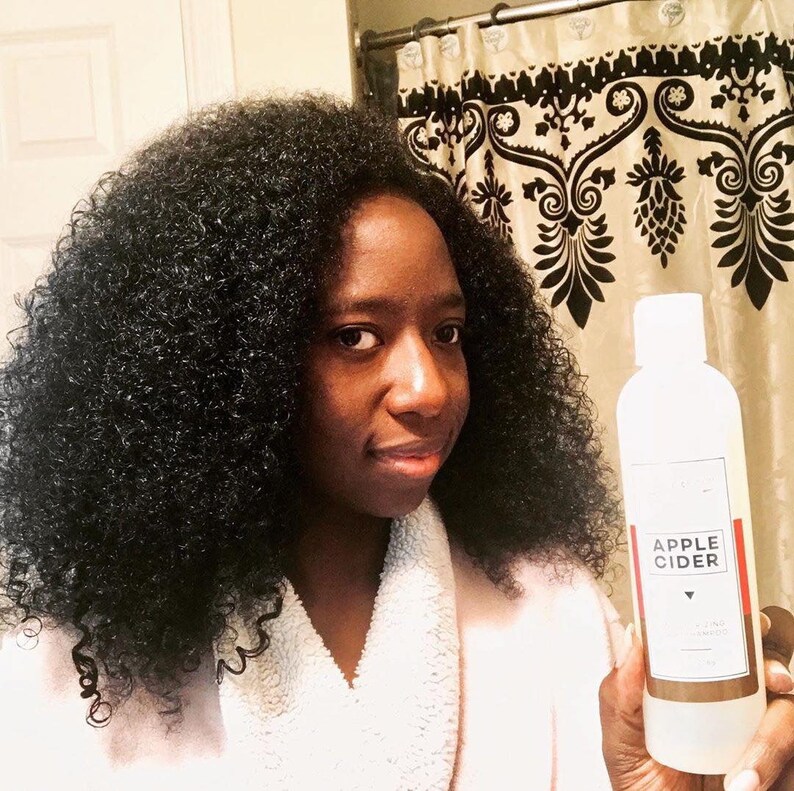 Puede incluir: Una persona con cabello negro rizado sostiene una botella transparente de champ&uacute; de vinagre de sidra de manzana. La botella tiene una etiqueta roja con las palabras "Apple Cider" en texto blanco.