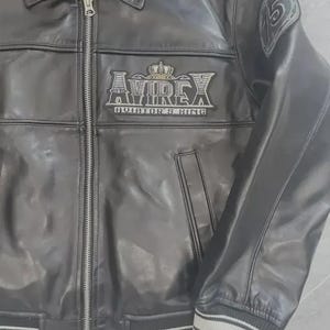 K&ouml;nnte beinhalten: Schwarze Lederjacke mit Rei&szlig;verschluss, Kragen und gerippten B&uuml;ndchen und Saum mit wei&szlig;en Streifen. Die Jacke hat das "AVIREX"-Logo mit Krone und dem Text "AVIATOR'S KING". Ein Aufn&auml;her mit der Zahl "5" befindet sich am Oberarm.