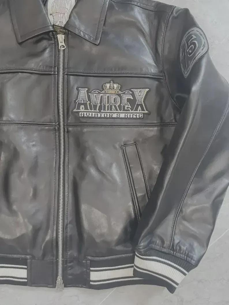 K&ouml;nnte beinhalten: Schwarze Lederjacke mit Rei&szlig;verschluss, Kragen und gerippten B&uuml;ndchen und Saum mit wei&szlig;en Streifen. Die Jacke hat das "AVIREX"-Logo mit Krone und dem Text "AVIATOR'S KING". Ein Aufn&auml;her mit der Zahl "5" befindet sich am Oberarm.