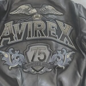 K&ouml;nnte beinhalten: Schwarze Lederjacke mit dem Wort "AVIREX" in gro&szlig;en Buchstaben auf dem R&uuml;cken. &Uuml;ber dem Wort befindet sich ein Adler mit ausgebreiteten Fl&uuml;geln. Darunter ein Schild mit der Zahl "75" und den Worten "AVIATOR'S KING".