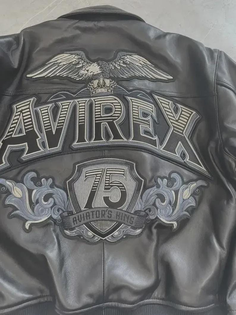K&ouml;nnte beinhalten: Schwarze Lederjacke mit dem Wort "AVIREX" in gro&szlig;en Buchstaben auf dem R&uuml;cken. &Uuml;ber dem Wort befindet sich ein Adler mit ausgebreiteten Fl&uuml;geln. Darunter ein Schild mit der Zahl "75" und den Worten "AVIATOR'S KING".
