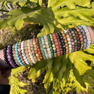 May include: A collection of beaded bracelets in various colors, including black, purple, turquoise, white, and shades of brown and green. The bracelets are displayed on a pink and gold cylindrical holder against a backdrop of large green leaves.