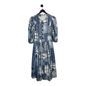 Può includere: Un abito vintage patchwork blu e bianco con bottoni sul davanti e maniche a tre quarti. L'abito è appeso a una gruccia di legno su sfondo bianco. L'abito presenta una varietà di motivi geometrici e floreali.