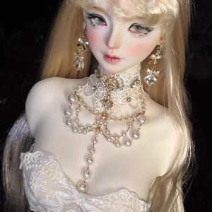 May include: A doll with long blonde hair, wearing a white lace choker necklace with gold accents and a pearl and gold chain. The doll also has matching earrings and a white lace dress.
