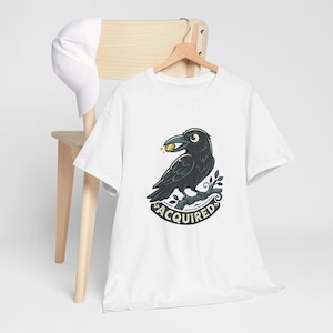 Pode incluir: Uma t-shirt branca de manga curta com um gráfico de um corvo preto pousado num ramo, segurando uma moeda de ouro no bico. Abaixo do corvo, a palavra ACQUIRED está impressa em letras escuras e negrito. Um boné branco simples também é visível.