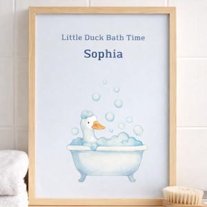 Könnte beinhalten: Ein gerahmter Druck mit einer Aquarellillustration einer weißen Ente in einer Badewanne mit Blasen. Der Text "Little Duck Bath Time" und der Name "Sophia" sind über der Ente abgebildet. Der Rahmen ist hellbraun.