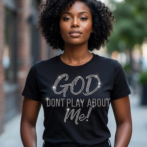 May include: Black t-shirt with the text "GOD DON'T PLAY ABOUT ME!" in sparkling rhinestones. The shirt is a crew neck style and has short sleeves. The text is in a script font.