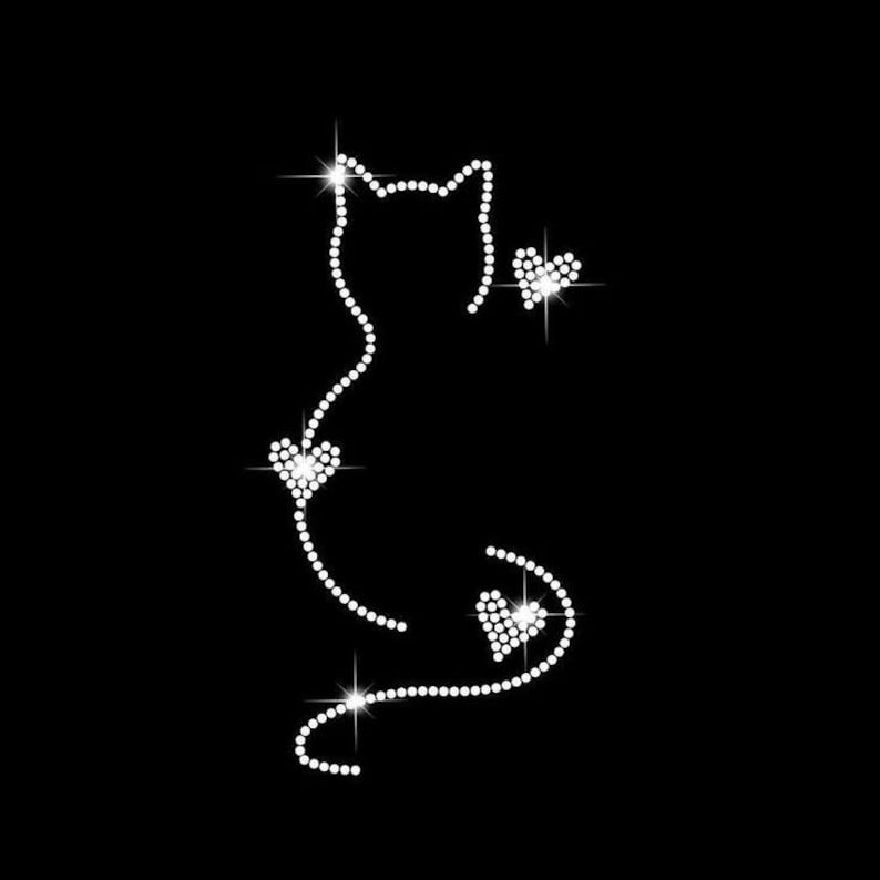 K&ouml;nnte beinhalten: Eine Katzen-Silhouette aus glitzernden Strasssteinen mit drei herzf&ouml;rmigen Akzenten. Der K&ouml;rper und der Schwanz der Katze werden durch eine Reihe kleiner, funkelnder Strasssteine gebildet. Die Herzen sind ebenfalls aus Strass, vor schwarzem Hintergrund.