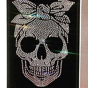 K&ouml;nnte beinhalten: Eine schwarze Leinwand zeigt ein Totenkopf-Design aus funkelnden Strasssteinen. Der Totenkopf hat eine Schleife im Bandana-Stil. Die Strasssteine sind so angeordnet, dass sie den Totenkopf und die Schleife bilden und dem Design einen Hauch von Glamour verleihen.