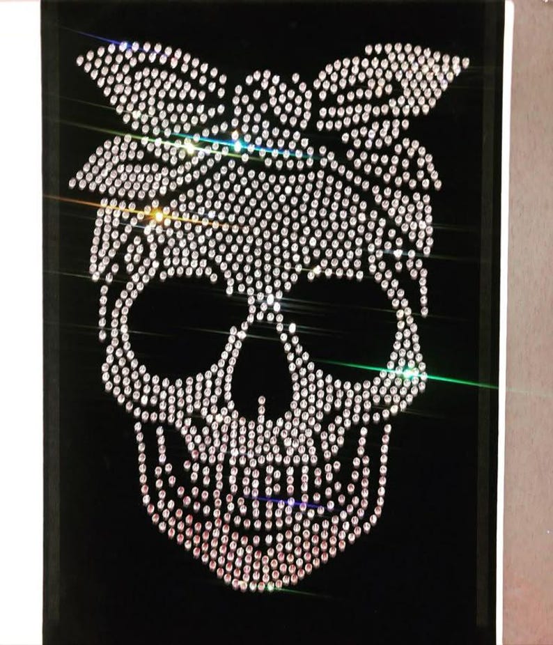 K&ouml;nnte beinhalten: Eine schwarze Leinwand zeigt ein Totenkopf-Design aus funkelnden Strasssteinen. Der Totenkopf hat eine Schleife im Bandana-Stil. Die Strasssteine sind so angeordnet, dass sie den Totenkopf und die Schleife bilden und dem Design einen Hauch von Glamour verleihen.