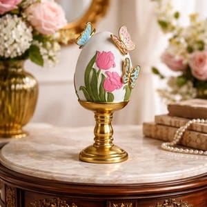 Può includere: Un ornamento decorativo a forma di uovo bianco con tulipani rosa e foglie verdi, adornato con farfalle dorate. L'uovo poggia su un piedistallo dorato, creando un'elegante esposizione a tema pasquale o primaverile.
