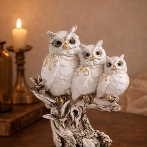 May include: A white owl family figurine perched on a detailed tree branch. The owls have gold accents and are arranged in a row. The sculpture is on a dark wooden surface.