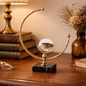May include: A decorative globe sculpture with a clear glass sphere held in a gold-coloured metal arc. It rests on a black marble base. The background includes a lamp, books, and dried flowers, creating a vintage aesthetic.