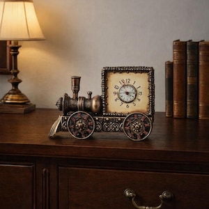 May include: A decorative antique-style clock shaped like a train. The clock has a square face with Roman numerals and a small round clock face. The train is bronze-colored with red accents on the wheels. The clock sits on a dark wooden surface.