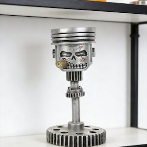 Puede incluir: Una escultura plateada única hecha con piezas de motor recicladas. Presenta una cabeza de pistón con forma de calavera con un cigarro, montada sobre una base en forma de engranaje. La escultura se exhibe en una estantería blanca.