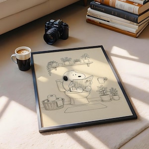 Puede incluir: Impresi&oacute;n art&iacute;stica en blanco y negro enmarcada de Snoopy. Snoopy est&aacute; sentado en un inodoro leyendo un peri&oacute;dico. La impresi&oacute;n art&iacute;stica est&aacute; rodeada de plantas en macetas y un rollo de papel higi&eacute;nico. Una c&aacute;mara y una taza de caf&eacute; tambi&eacute;n est&aacute;n en la imagen.