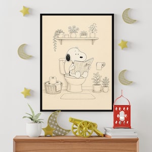 Puede incluir: Ilustraci&oacute;n en blanco y negro enmarcada de Snoopy sentado en un inodoro, leyendo un peri&oacute;dico. La escena del ba&ntilde;o incluye una estanter&iacute;a con plantas en macetas, un rollo de papel higi&eacute;nico y una cesta tejida con rollos adicionales. La obra de arte se exhibe en una pared con estrellas y lunas decorativas.