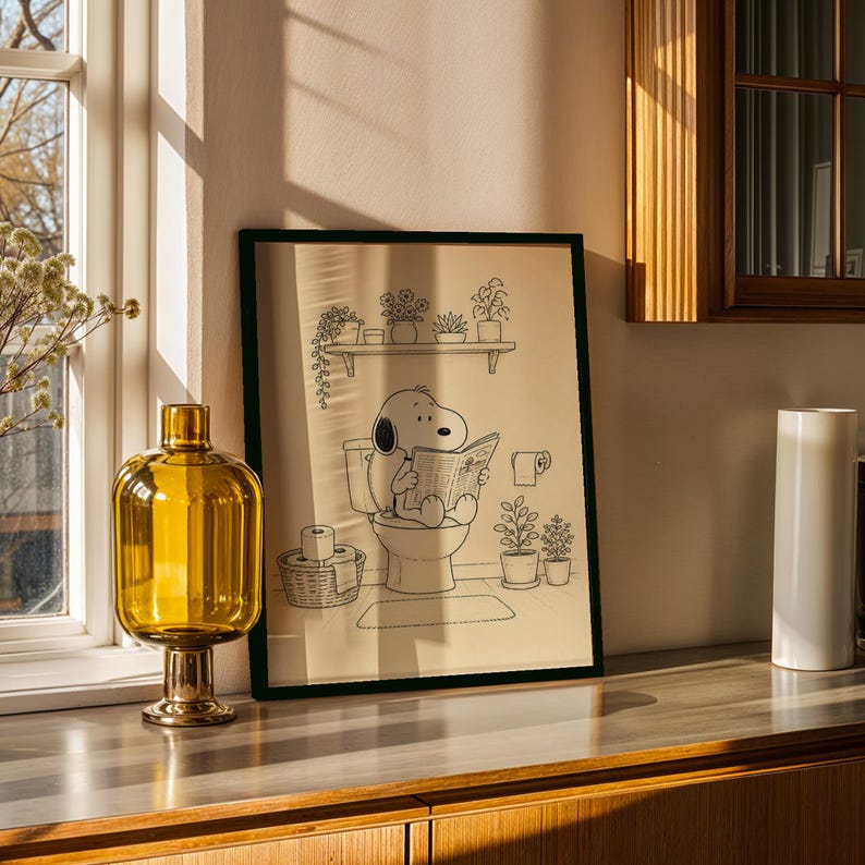 Puede incluir: Ilustraci&oacute;n en blanco y negro enmarcada de Snoopy leyendo un peri&oacute;dico en el inodoro. La obra de arte se exhibe en una encimera con un jarr&oacute;n de cristal amarillo y un objeto cil&iacute;ndrico blanco.