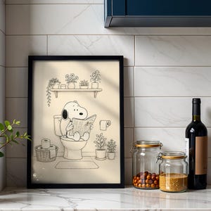Puede incluir: Ilustraci&oacute;n en blanco y negro enmarcada de Snoopy leyendo un peri&oacute;dico en un inodoro. La obra de arte se presenta en un ba&ntilde;o, con una estanter&iacute;a con plantas encima y un rollo de papel higi&eacute;nico.