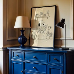 Puede incluir: Un estampado en blanco y negro enmarcado de Snoopy en el inodoro leyendo un peri&oacute;dico. El estampado se muestra en una c&oacute;moda azul, junto a una l&aacute;mpara azul con una pantalla crema y una l&aacute;mpara de escritorio negra.