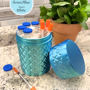 May include: A cylindrical green and blue container with a textured diamond pattern, featuring a matching lid, a white interior insert, and several small glass vials with blue caps and syringes with orange caps stored inside and placed nearby.