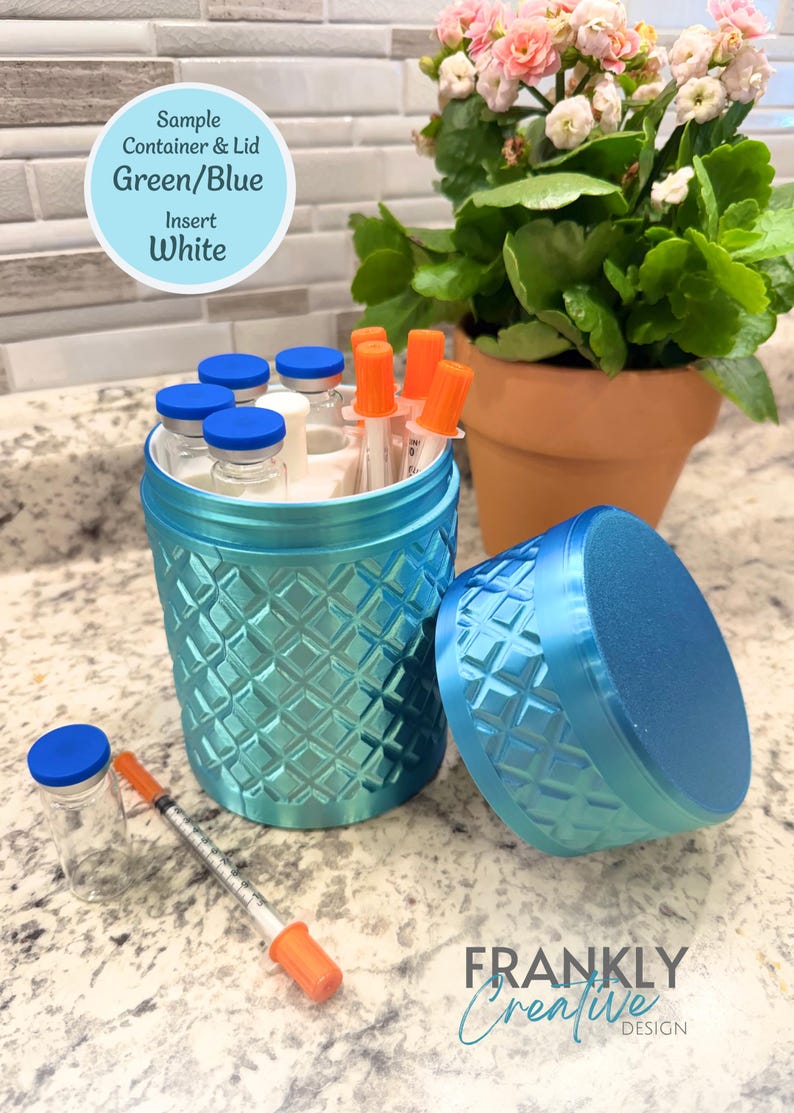 May include: A cylindrical green and blue container with a textured diamond pattern, featuring a matching lid, a white interior insert, and several small glass vials with blue caps and syringes with orange caps stored inside and placed nearby.