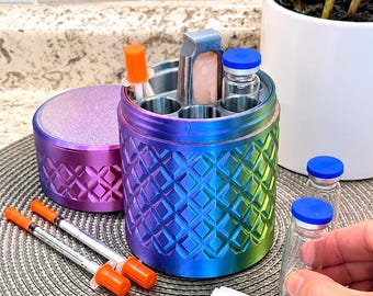 May include: A cylindrical, iridescent metal storage container with a diamond-patterned exterior, featuring internal slots holding small glass vials with blue caps and an orange-capped syringe. Includes two additional syringes and an alcohol prep pad.