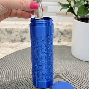 May include: A cylindrical blue container with a textured surface featuring a repeating pattern of small, rounded triangular shapes. The container has a matching blue screw-on lid and an internal component being inserted into the top opening.