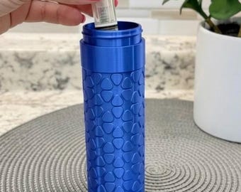 May include: A cylindrical blue container with a textured surface featuring a repeating pattern of small, rounded triangular shapes. The container has a matching blue screw-on lid and an internal component being inserted into the top opening.