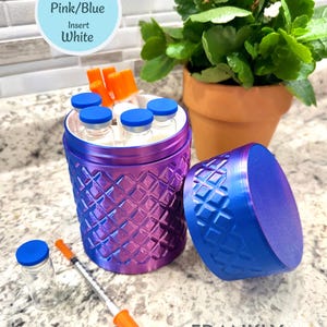 May include: A cylindrical container with a pink and blue gradient diamond-textured exterior and a matching lid. The container holds several small glass vials with blue caps and orange-capped syringes. Text reads: Sample Container & Lid Pink/Blue Insert White.