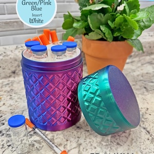 May include: A cylindrical container with a diamond-textured surface in a gradient of green, pink, and blue. It features a white interior insert, a matching lid, and holds several small glass vials with blue caps and orange-capped syringes.