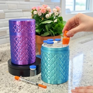 May include: A purple cylindrical container with a diamond-patterned texture and a matching blue container holding several small glass vials with blue and orange caps. A single syringe with an orange cap and a small glass vial sit on the counter.