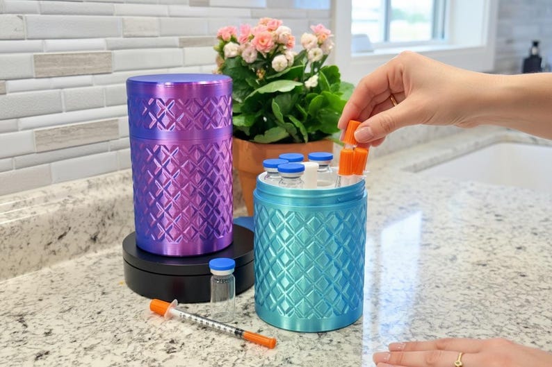 May include: A purple cylindrical container with a diamond-patterned texture and a matching blue container holding several small glass vials with blue and orange caps. A single syringe with an orange cap and a small glass vial sit on the counter.