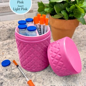 May include: A pink cylindrical container with a textured diamond pattern and a light pink insert, accompanied by a matching lid. The container holds several small glass vials with blue caps and syringes with orange caps, with one additional vial and syringe nearby.