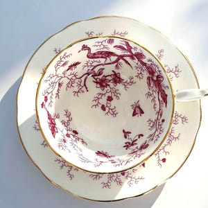 May include: A vintage teacup and saucer set. The white porcelain cup and saucer feature a burgundy bird and floral design. The rims are scalloped and trimmed with gold. A classic addition to any tea set collection.