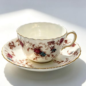 May include: A cream-coloured porcelain teacup and saucer set featuring a scalloped edge and a delicate floral pattern in shades of deep red, brown, and blue. The rim of both the cup and the saucer is finished with a thin, reflective gold trim.