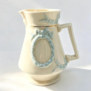 May include: A cream-coloured ceramic pitcher featuring a raised, light blue ribbon and bow motif in an oval shape on the front. The pitcher has a gold-toned rim accent, a wide spout, and a geometric handle, presenting a smooth, glossy finish.