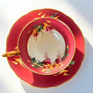 May include: A vintage teacup and saucer set in deep red with gold trim. The teacup and saucer are decorated with a floral design in red, yellow, and green. The teacup has a built-in cigar rest. A classic tea set.